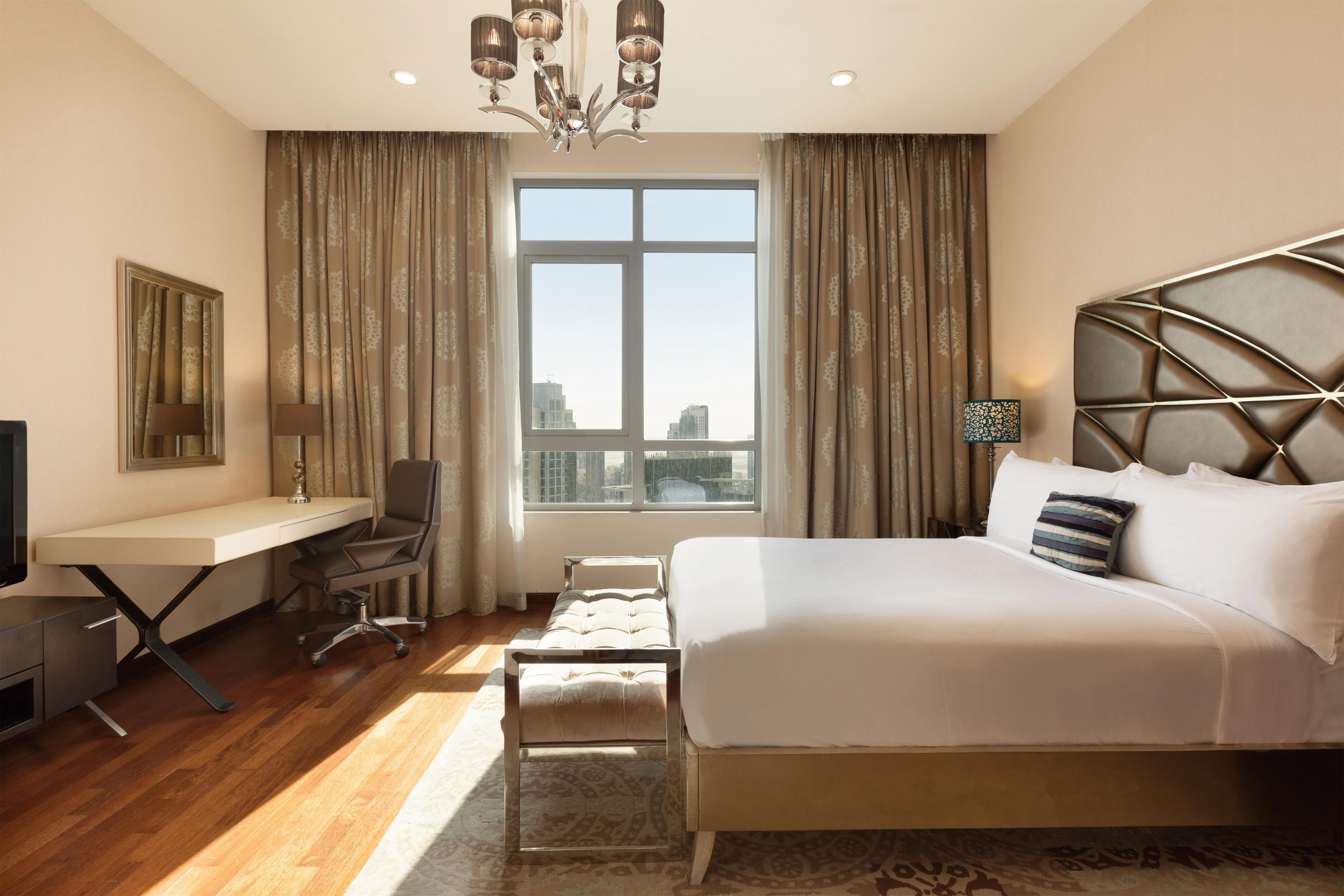 A hotel room with a bed, desk, chair, and a large window with a view of buildings.