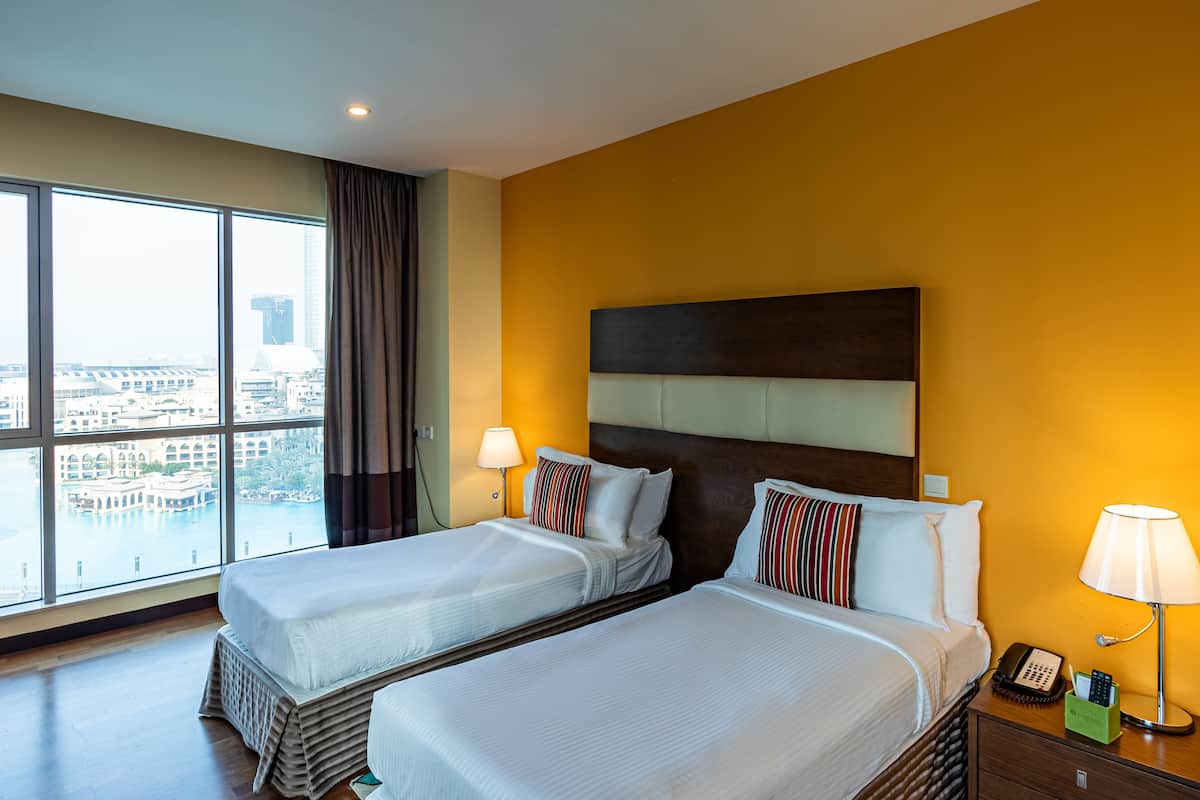 Een hotelkamer met twee bedden, een groot raam met uitzicht op de stad, een houten hoofdbord en een nachtkastje met een lamp en telefoon.