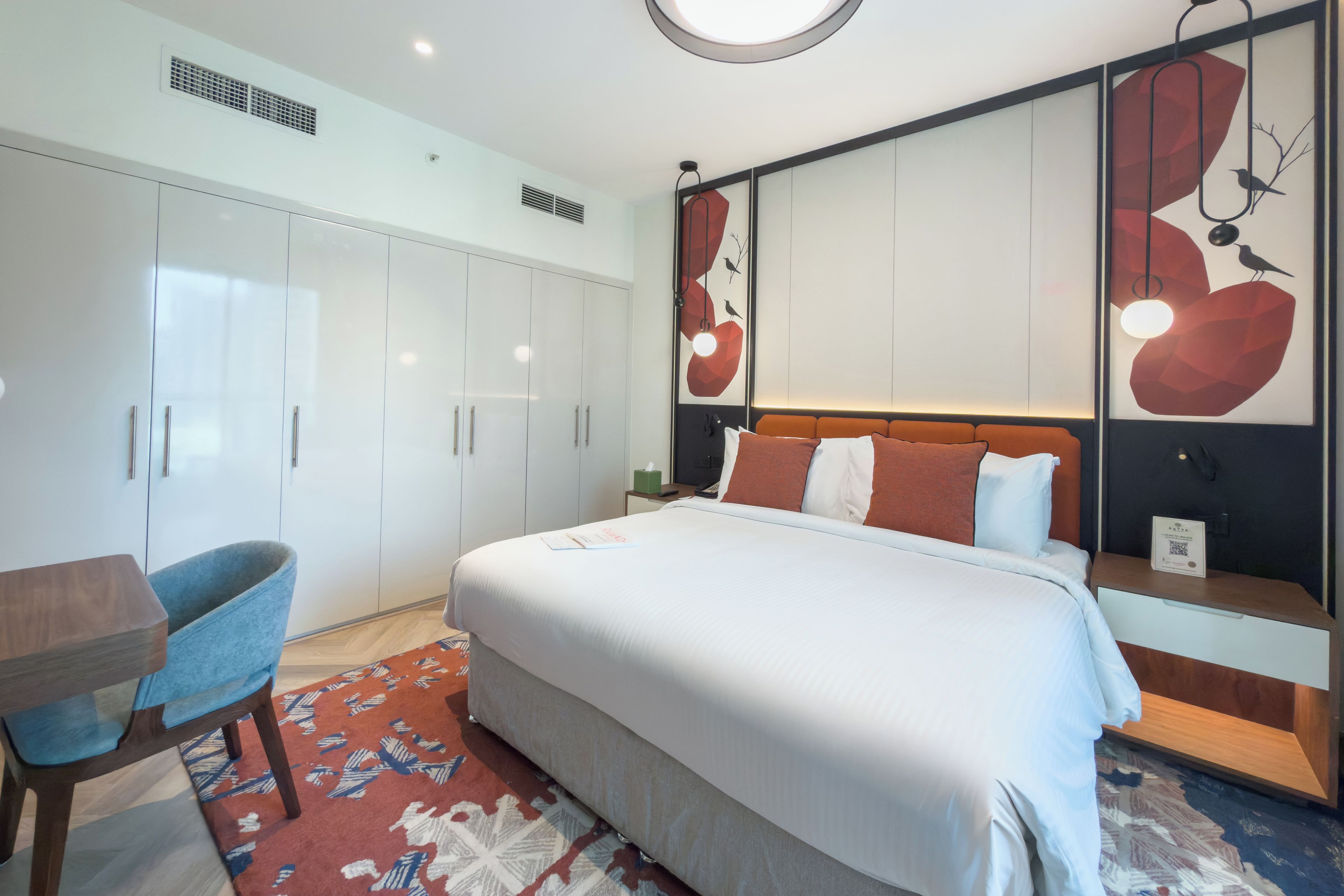 Ein modernes Hotelzimmer mit einem großen Bett, einem Schreibtisch und einem Stuhl. Es gibt Schränke, einen Nachttisch und eine dekorative Wandgestaltung.