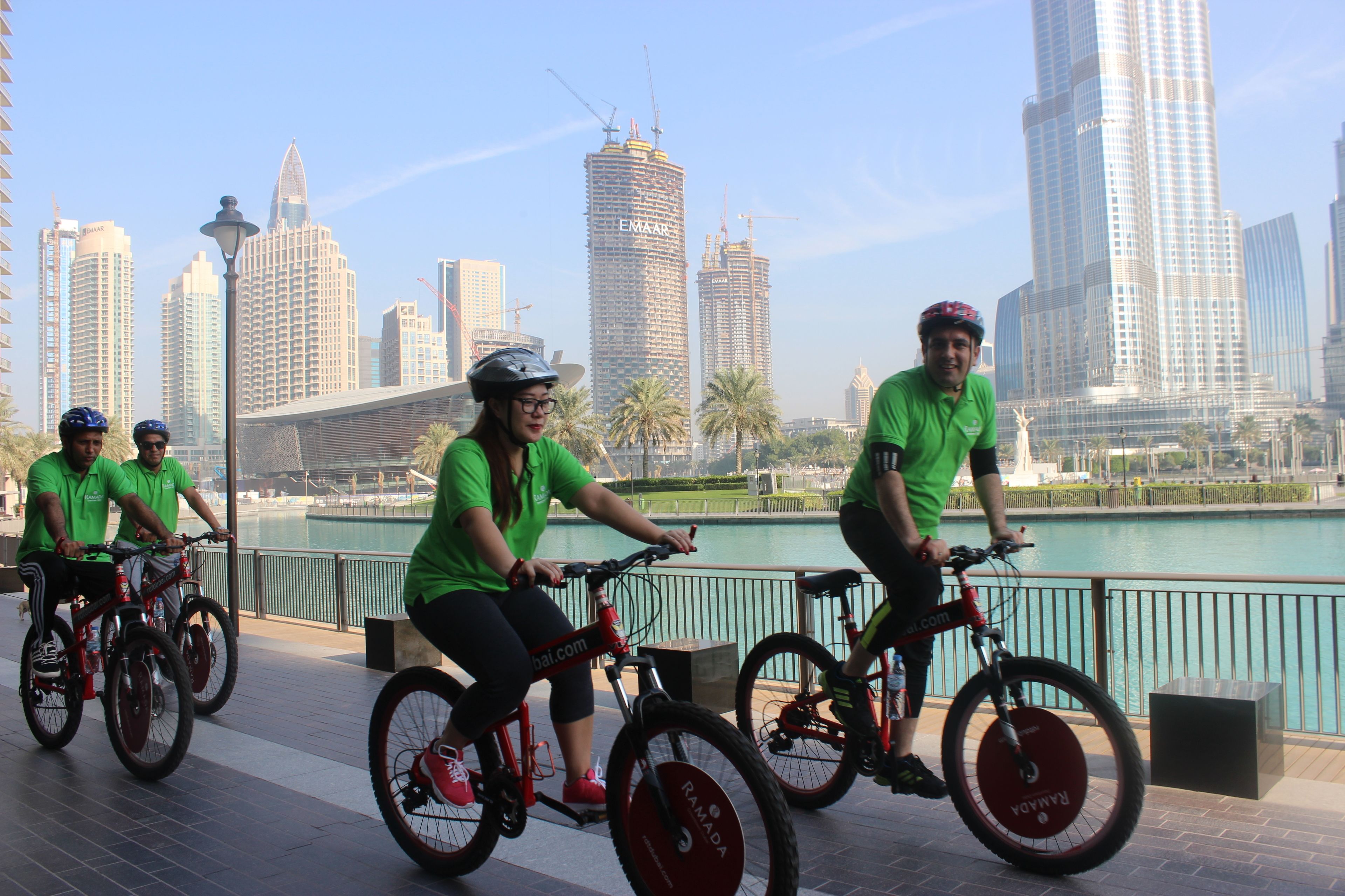 A group of people riding bicycles along a waterfront promenade with a city skyline in the background.