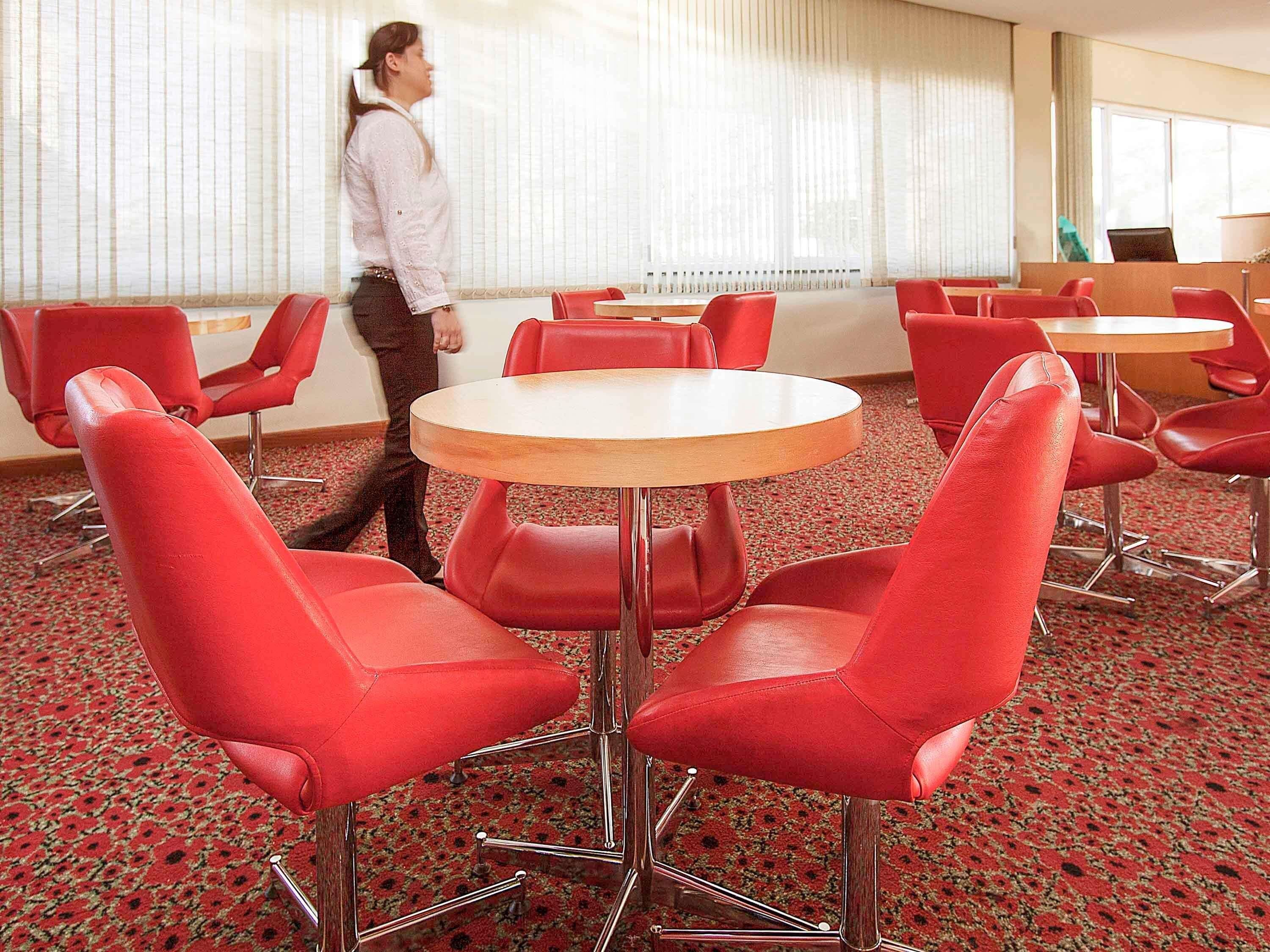 A hotel room with red chairs around a central table, a large window with blinds, and a carpeted floor.