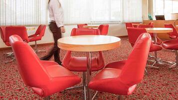 A hotel room with red chairs around a central table, a large window with blinds, and a carpeted floor.