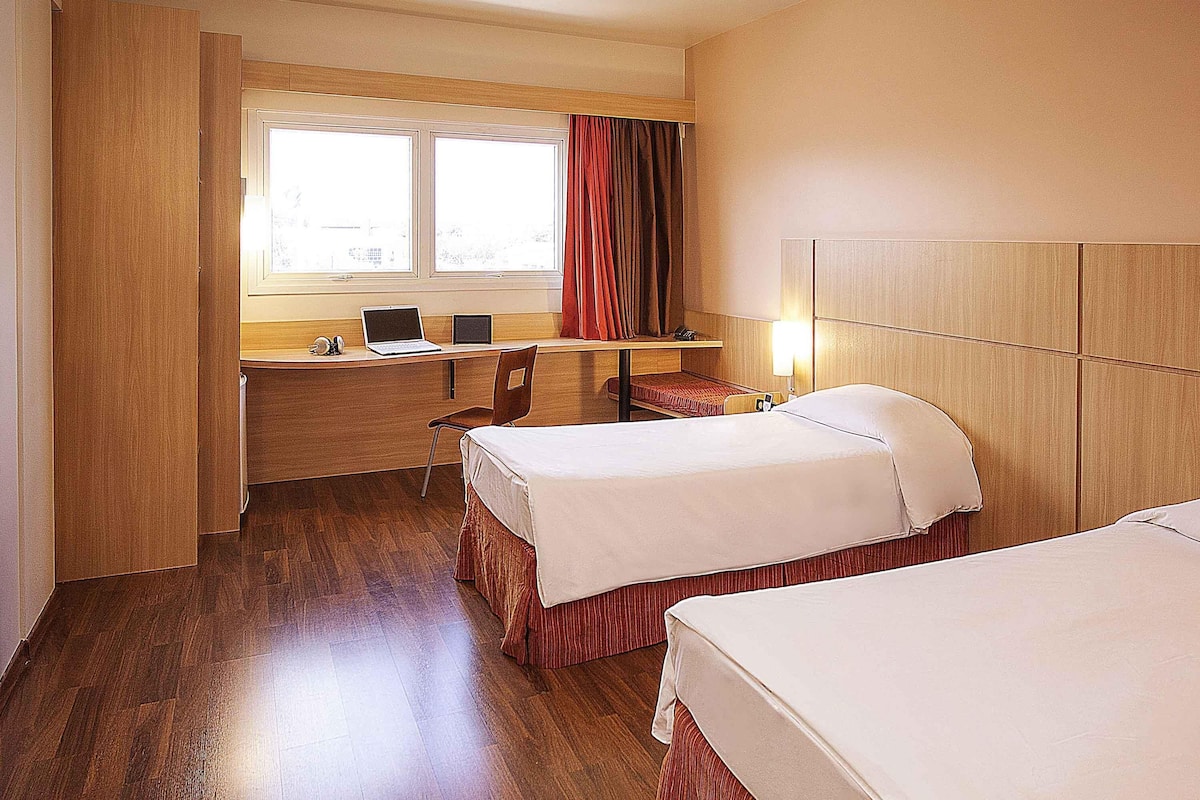 A hotel room with two beds, a desk with a laptop, a chair, and a window with curtains.