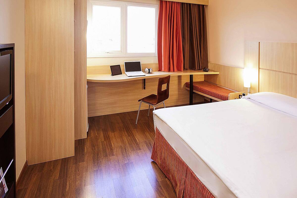 A hotel room with a wooden desk, a laptop, a chair, a bed, and a television.