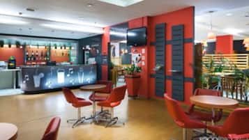 Un café moderno con sillas y mesas rojas, una zona de bar con mostrador, y un televisor de pantalla plana montado en la pared.