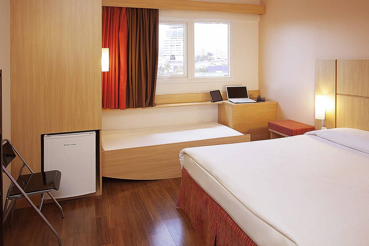 A hotel room with a bed, a desk with a laptop, a small fridge, and a window with curtains.