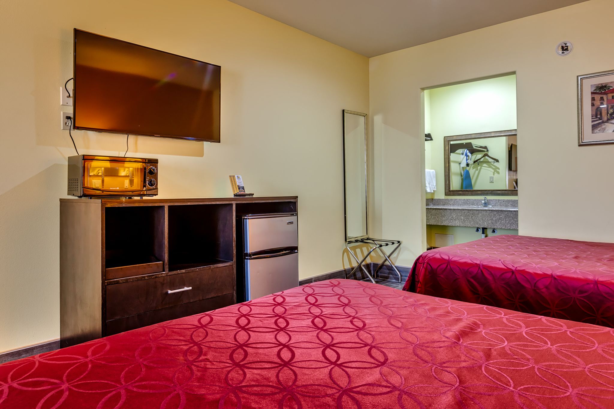 Habitación de hotel con televisor montado en la pared, microondas, refrigerador, espejo y dos camas con ropa de cama roja.