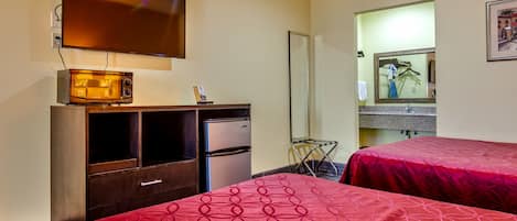 Habitación de hotel con televisor montado en la pared, microondas, refrigerador, espejo y dos camas con ropa de cama roja.