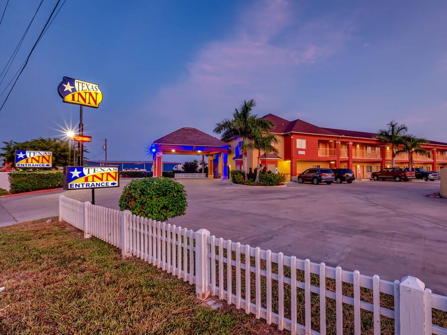 A Texas Inn hotel with a well-lit entrance, a parking lot, and a white picket fence.