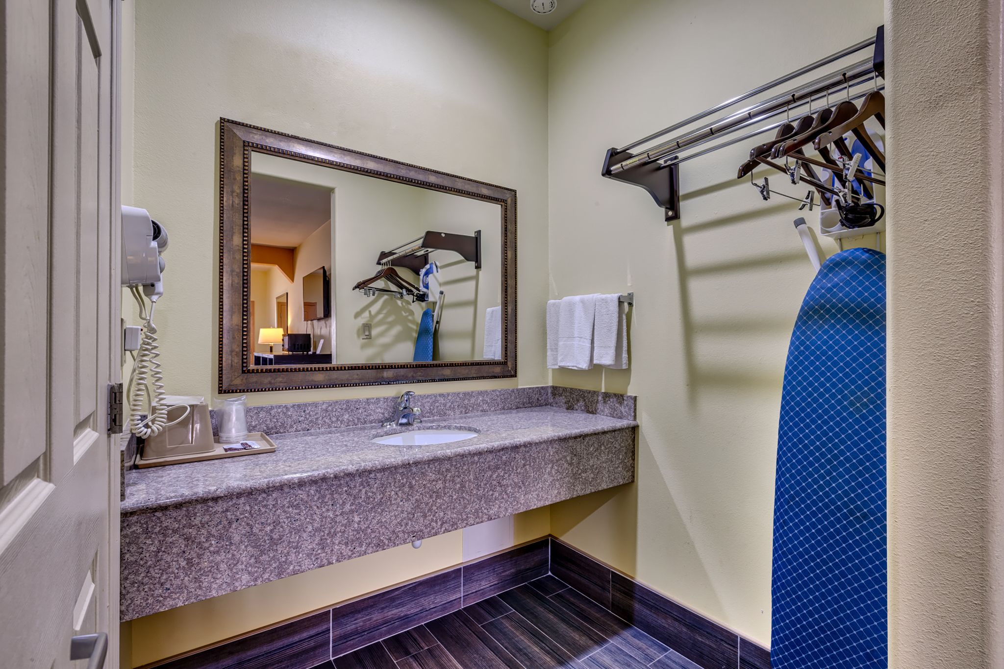 Un baño de hotel con lavamanos de encimera de granito, un espejo grande, un secador de pelo y un perchero para toallas.