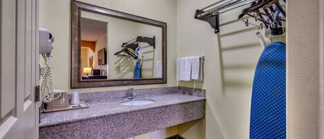Un baño de hotel con lavamanos de encimera de granito, un espejo grande, un secador de pelo y un perchero para toallas.