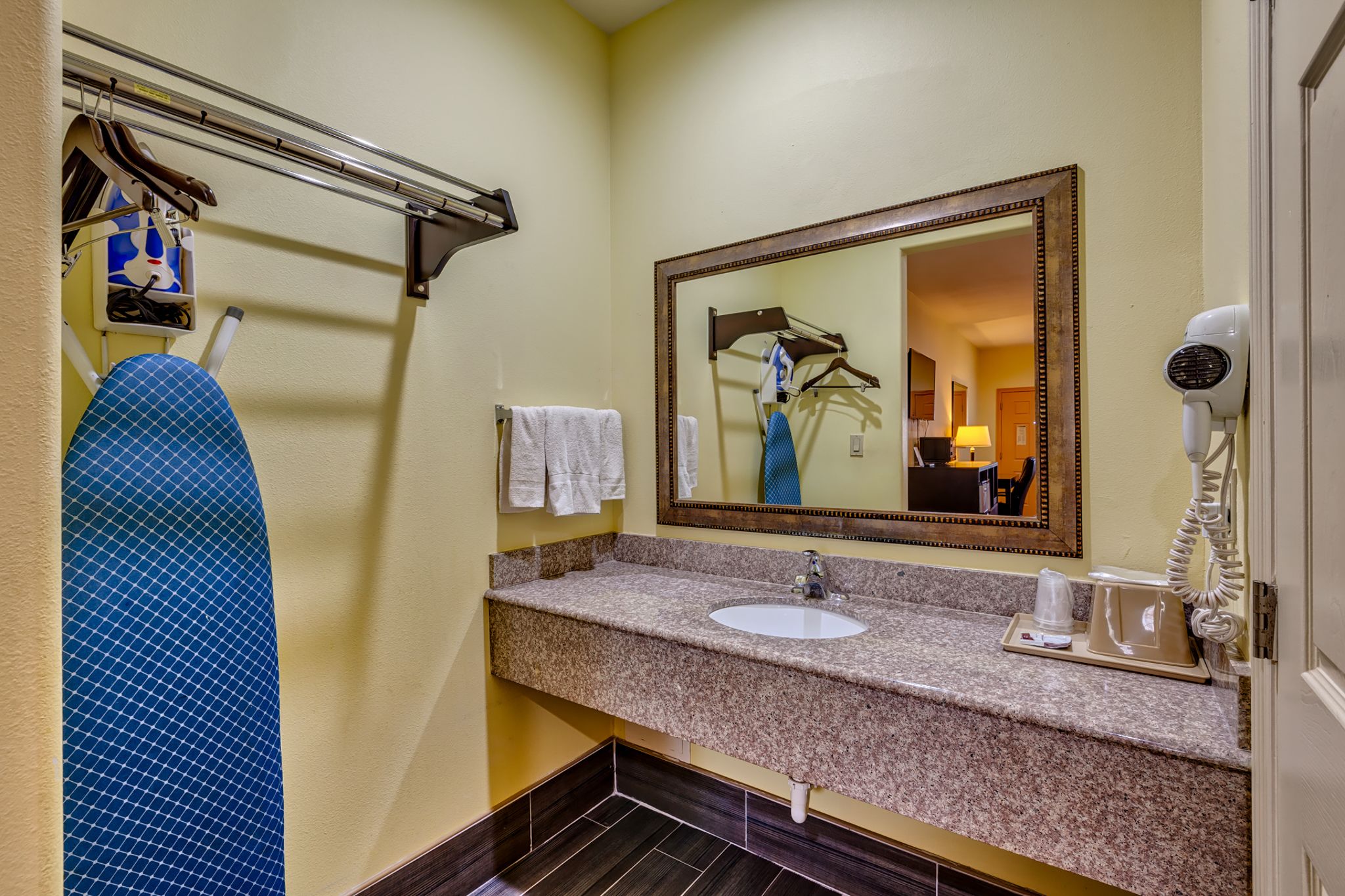 A bathroom with a granite countertop, a sink, a mirror, a towel rack, a hairdryer, and a shower chair.