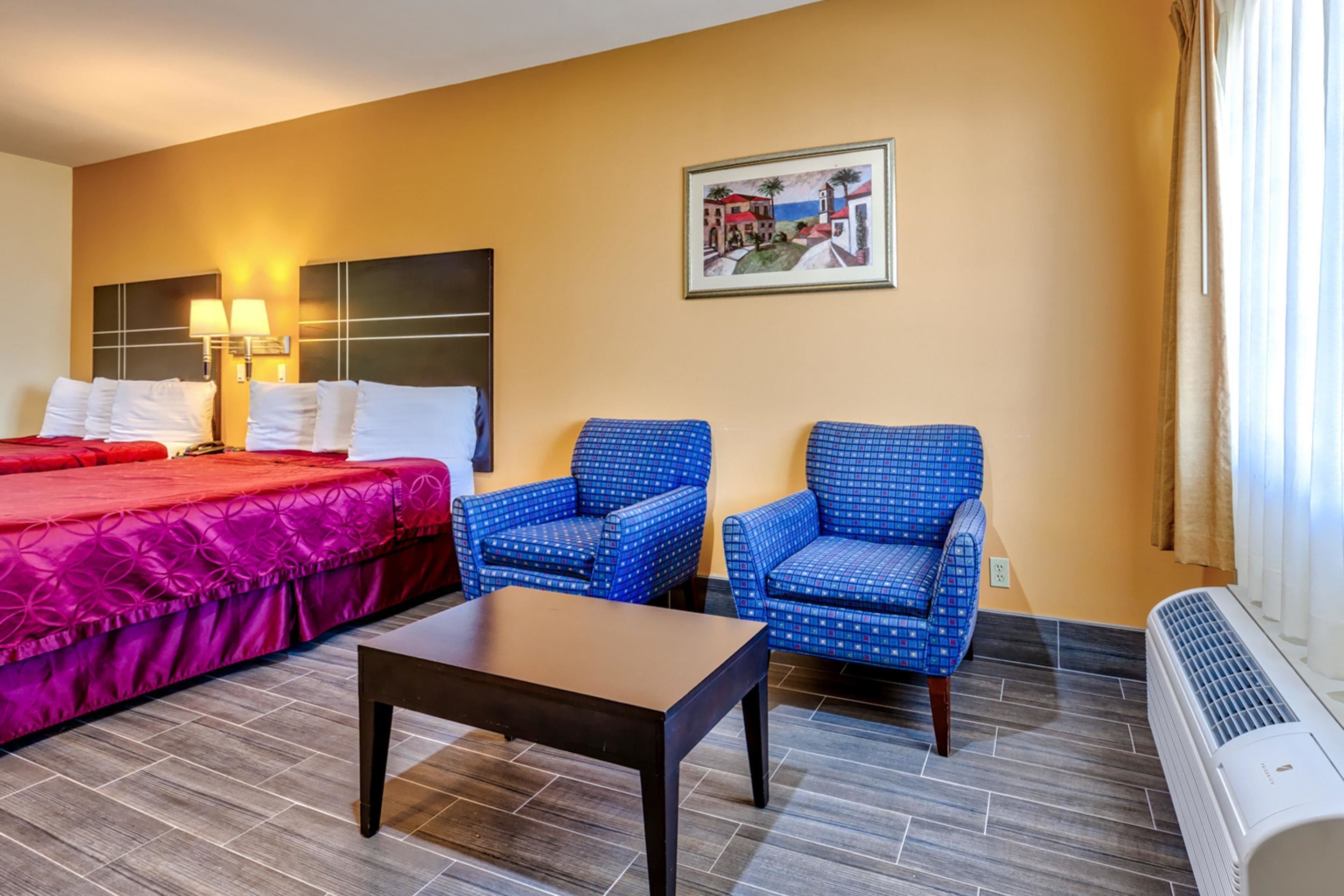 Habitación de hotel con una cama, dos sillas azules con estampado, una mesita pequeña, un calentador de pared y un cuadro de una escena costera.