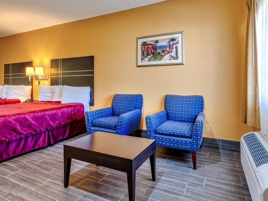 Habitación de hotel con una cama, dos sillas azules con estampado, una mesita pequeña, un calentador de pared y un cuadro de una escena costera.