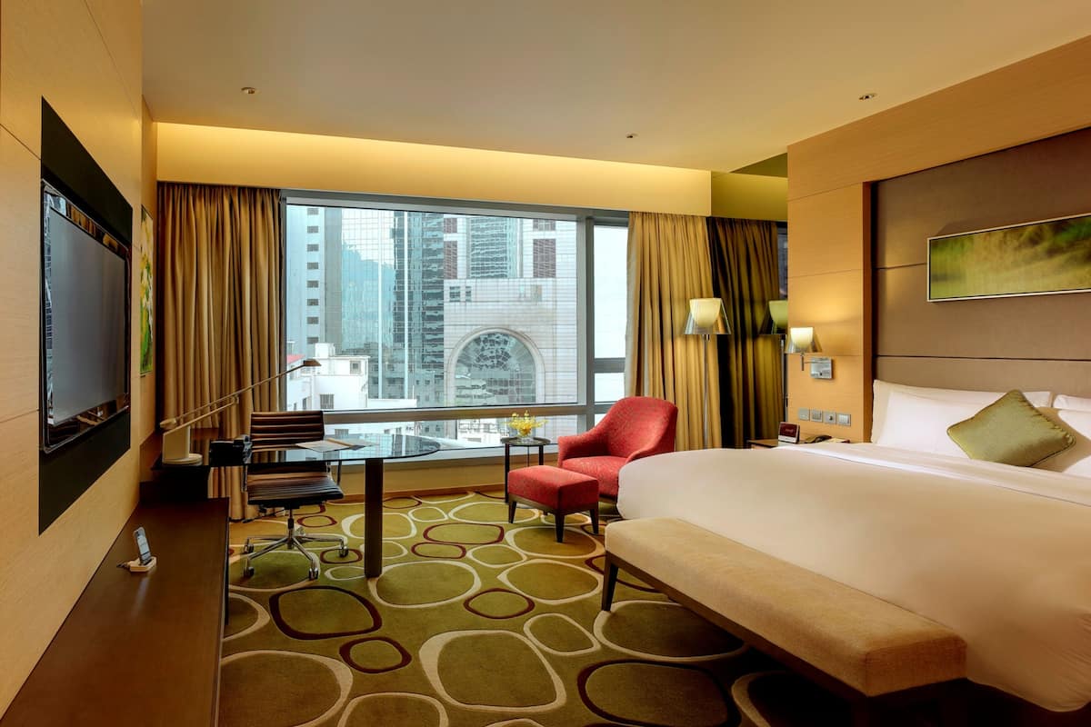 A hotel room with a large bed, a desk with a chair, a TV, and a view of a cityscape through large windows.