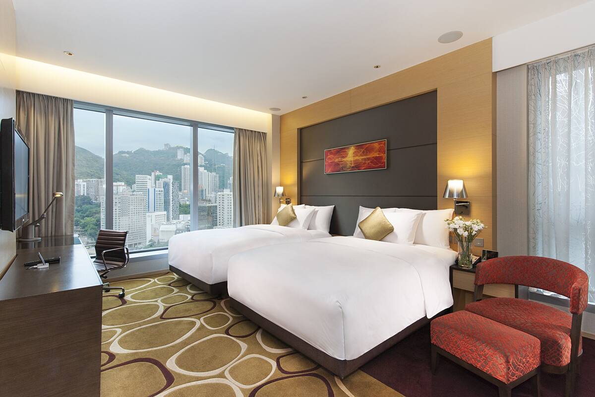 A hotel room with two beds, a desk, a chair, and a large window offering a city view.
