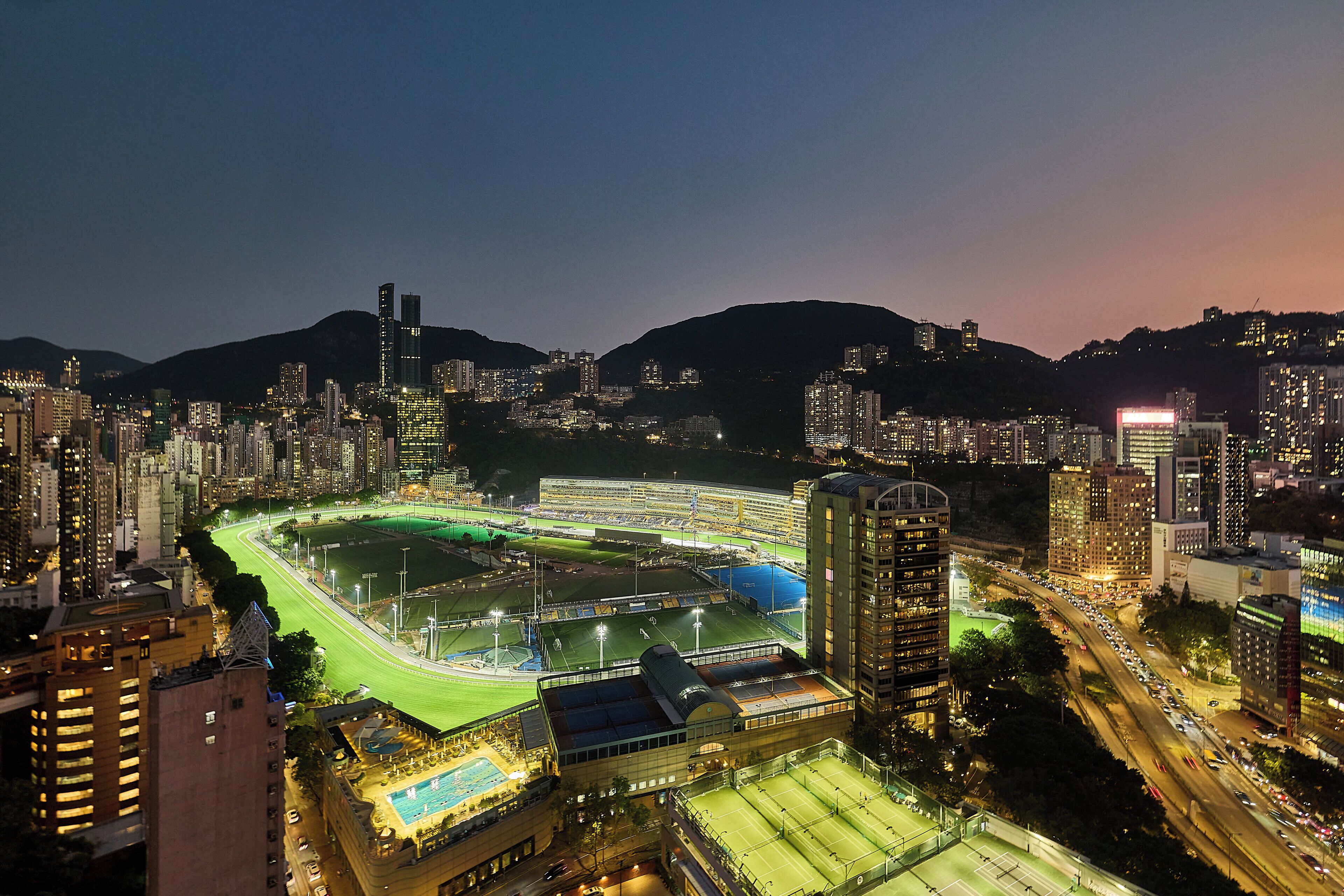 A cityscape at night with illuminated buildings, a sports complex, and a road.