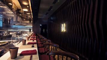 A restaurant with a long counter, multiple chairs, and a kitchen in the background.