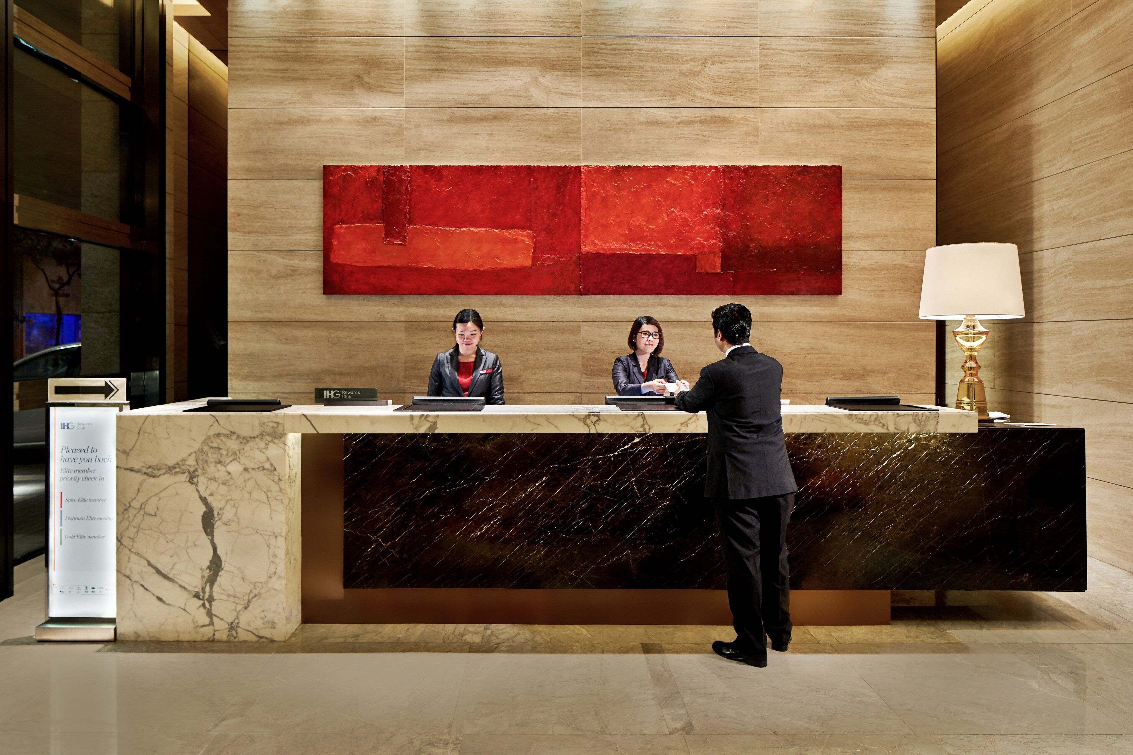 Un mostrador de recepción de hotel con tres empleados, un cuadro abstracto grande y rojo en la pared, y una lámpara moderna a la derecha.