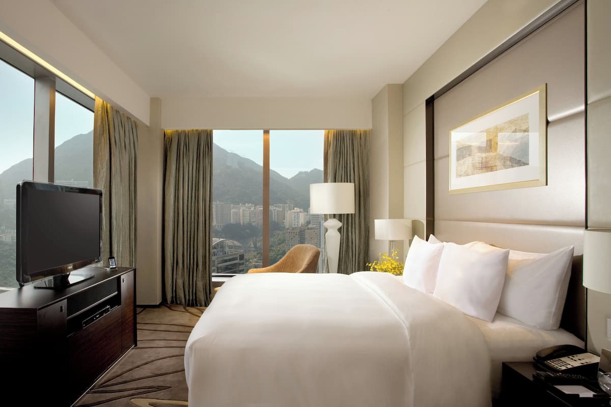 A hotel room with a large bed, a TV, a chair, and a desk. There is a view of mountains and buildings through the windows.