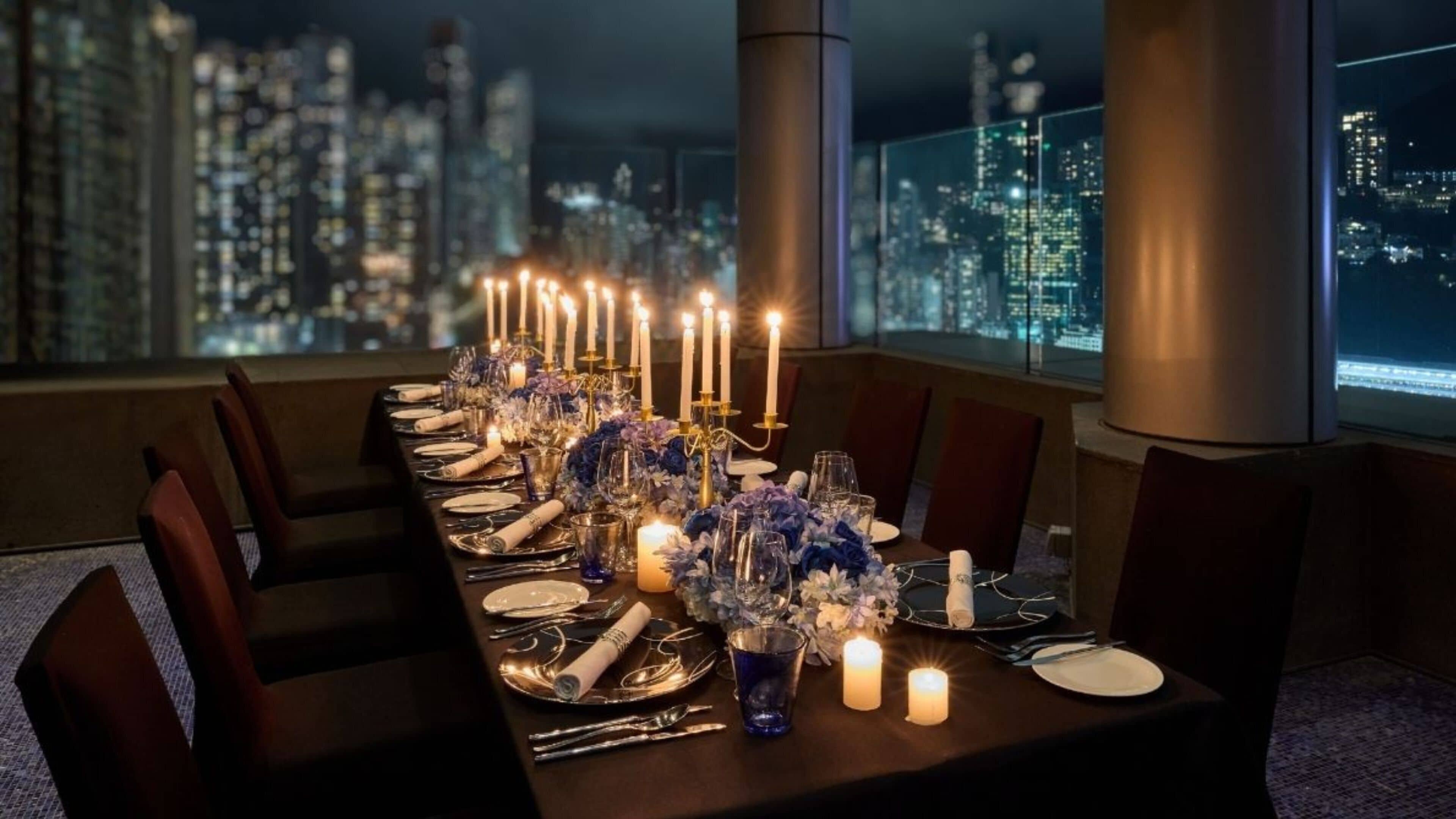 Ein langer Esstisch, gedeckt für eine Mahlzeit mit Kerzen, Gläsern und Besteck, mit Blick auf eine nächtliche Stadtlandschaft.