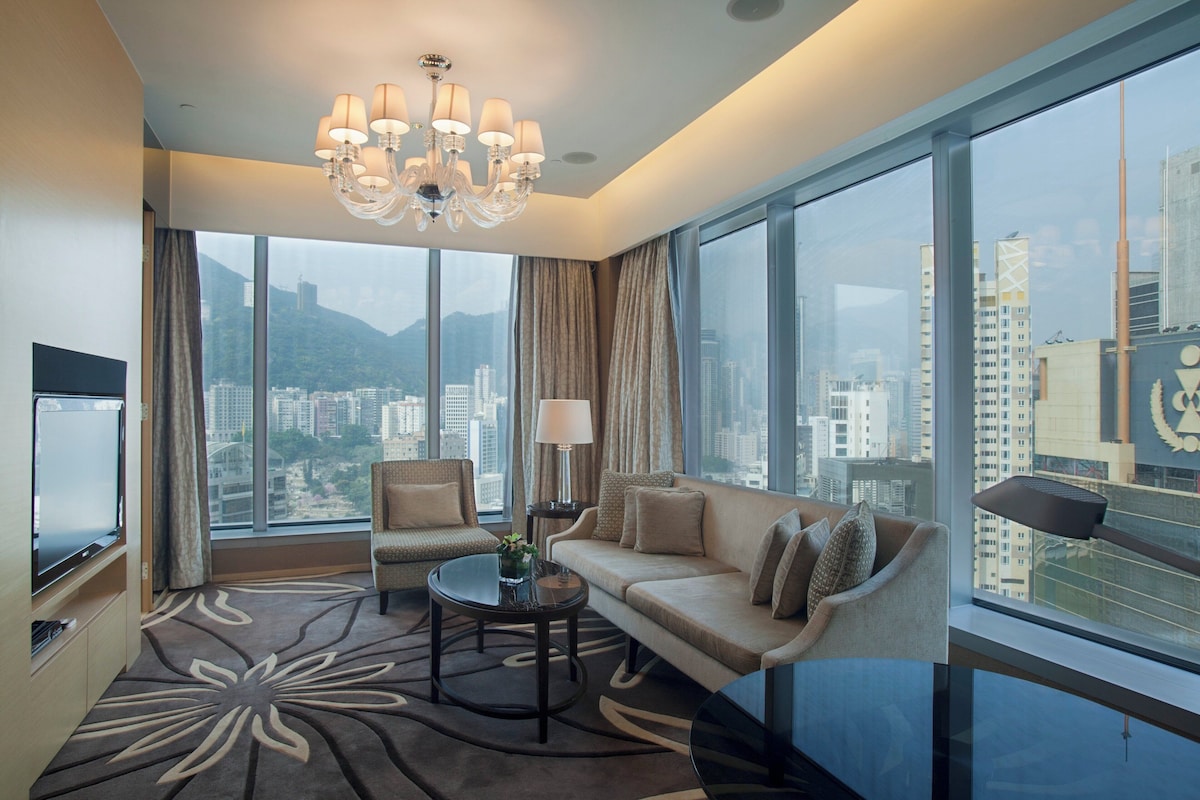 A modern hotel room with a large window, a sofa, a chair, a coffee table, and a TV.