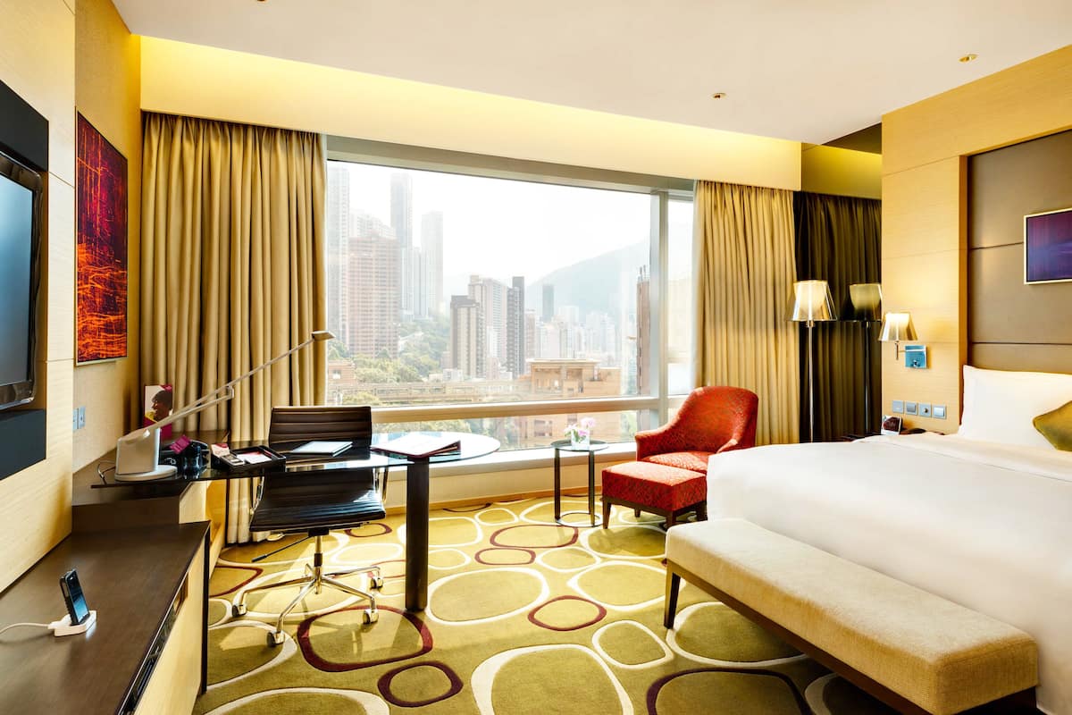 A hotel room with a large bed, a desk with a chair, a small table, and a red chair. There is a large window with a city view.