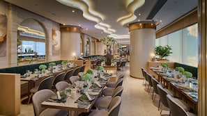 A restaurant with long communal tables, high ceilings with wavy lighting, and large windows.
