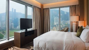 Habitación de hotel con una cama grande, un televisor sobre un mueble de madera y vistas a un paisaje urbano a través de amplias ventanas.