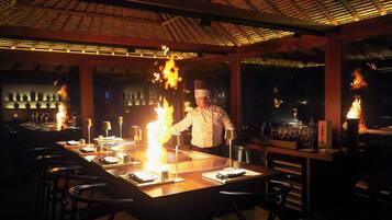 A chef preparing food at a table with flames, surrounded by bar stools and wine glasses.