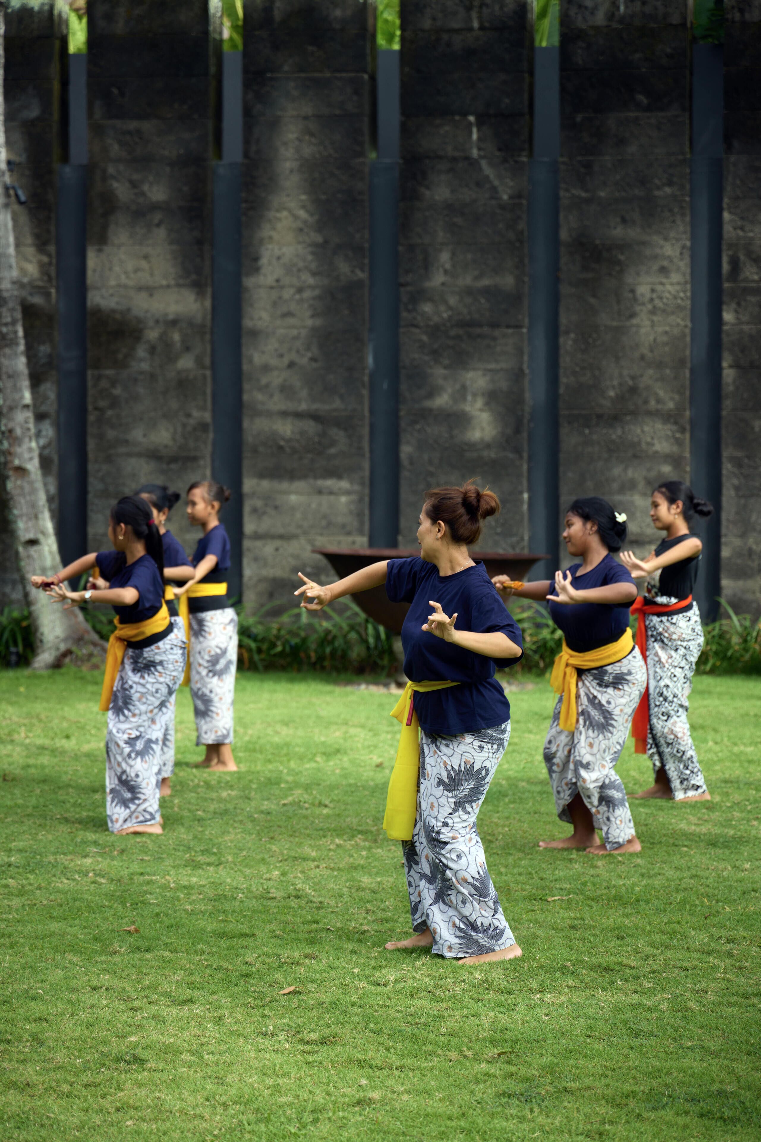 A group of people practicing martial arts outdoors on a grassy area with a stone wall in the background.