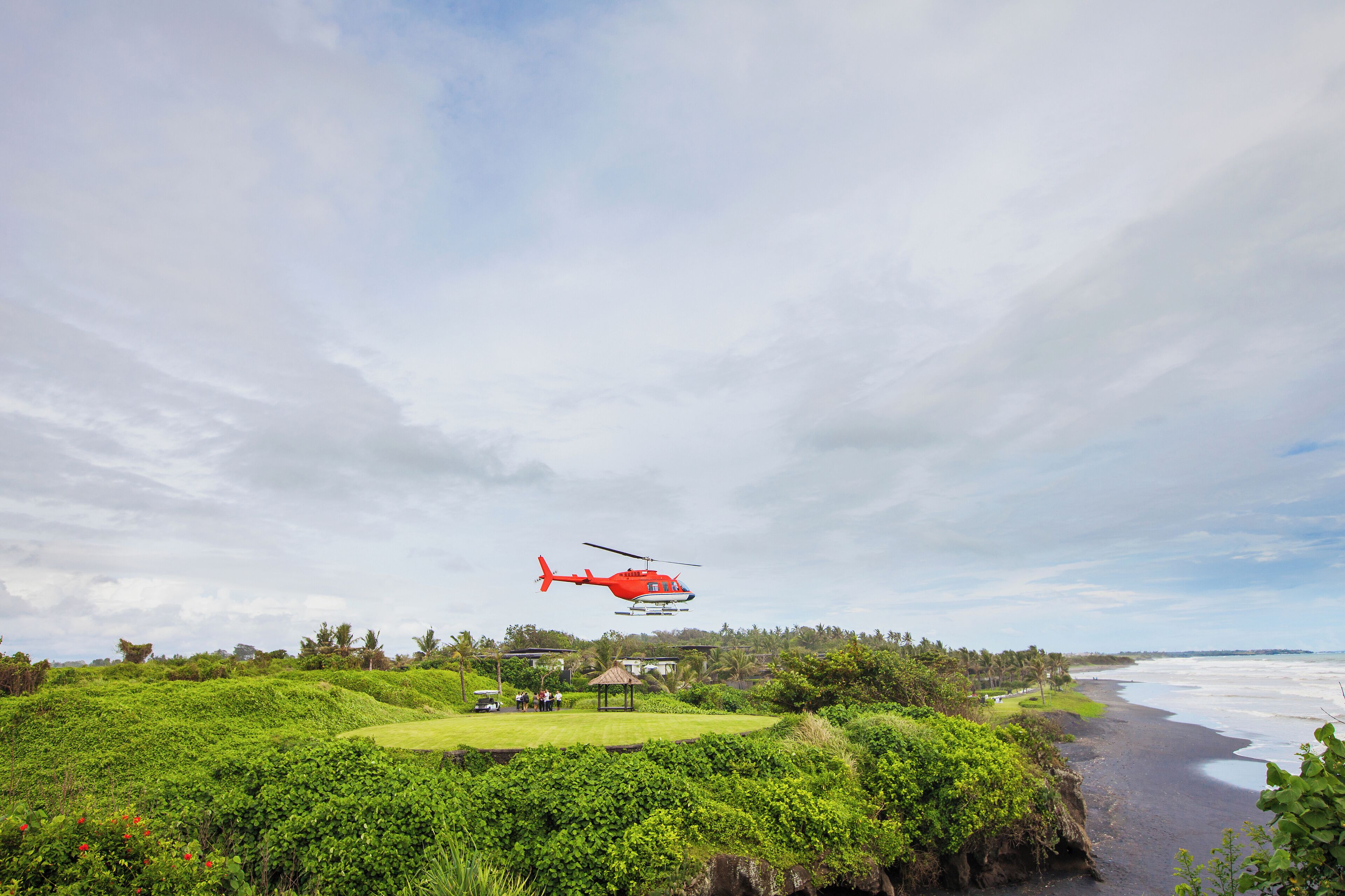 A red helicopter flying over a lush green landscape with a beach in the background.
