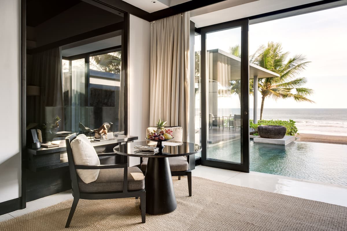 A modern living room with a glass wall overlooking a pool and beach, featuring a round table, chairs, and a view of the ocean.