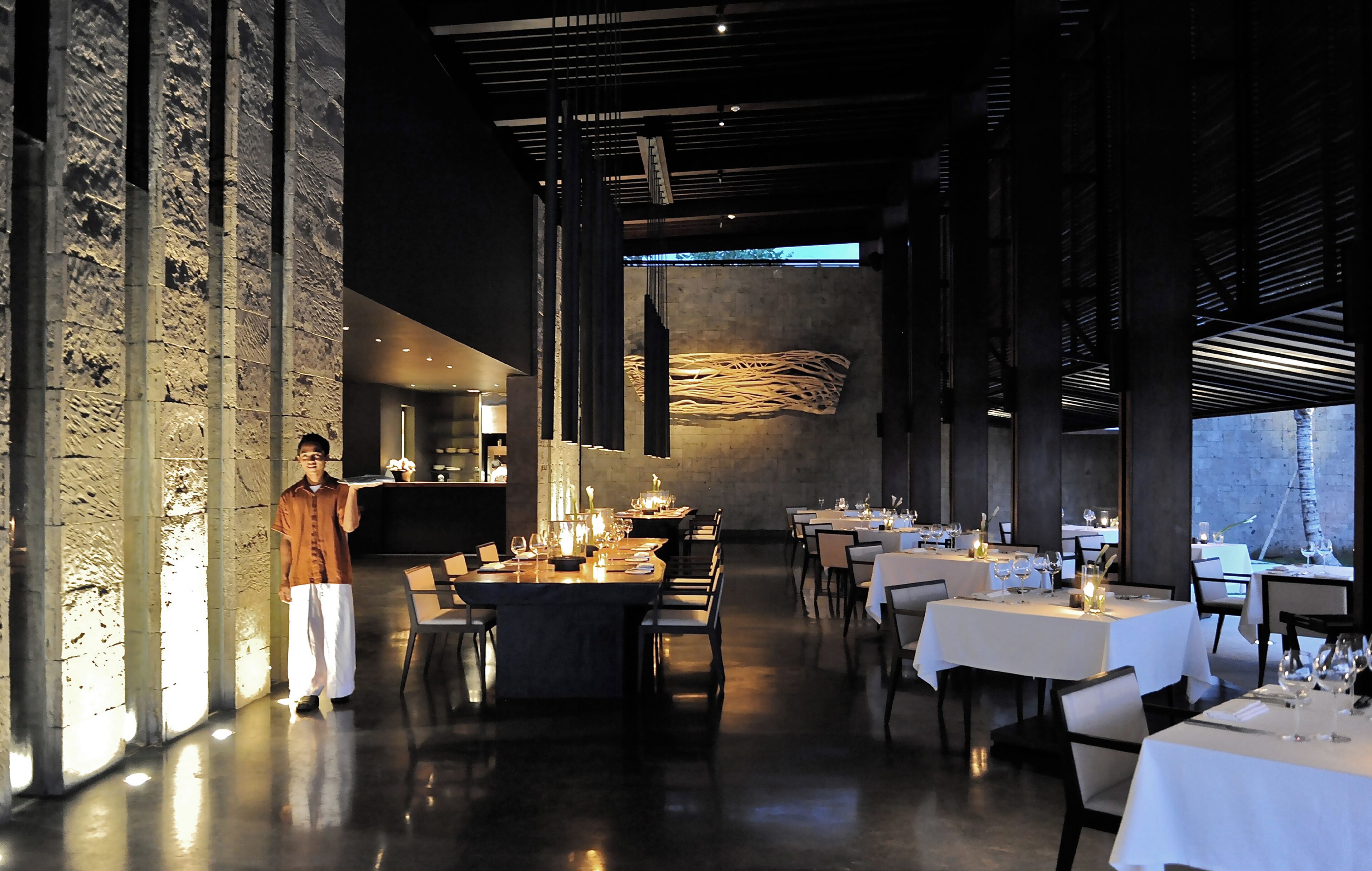A modern restaurant interior with high ceilings, exposed brick walls, and a long dining area with tables set for multiple guests.