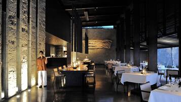 A modern restaurant interior with high ceilings, exposed brick walls, and a long dining area with tables set for multiple guests.