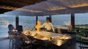 A restaurant with a large dining table, a chef performing a fire cooking technique, and a view of the sea. - Soori Bali (Tabanan)