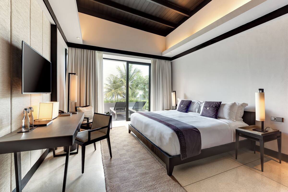 A modern hotel room with a large bed, a desk, a flat-screen TV, and a balcony with a view of palm trees.