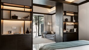 A modern bedroom with a large bed, built-in shelving with decorative items, a sofa, and a view of palm trees through a glass door. - Soori Bali (Tabanan)
