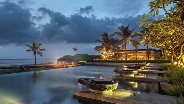 A serene evening pool area with a traditional water feature, palm trees, and a well-lit building in the background. - Soori Bali (Tabanan)