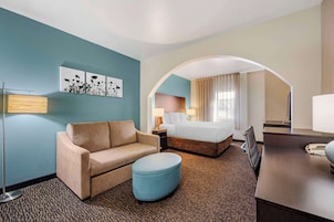 A hotel room with a sofa, ottoman, bed, desk, lamp, and artwork on the wall.