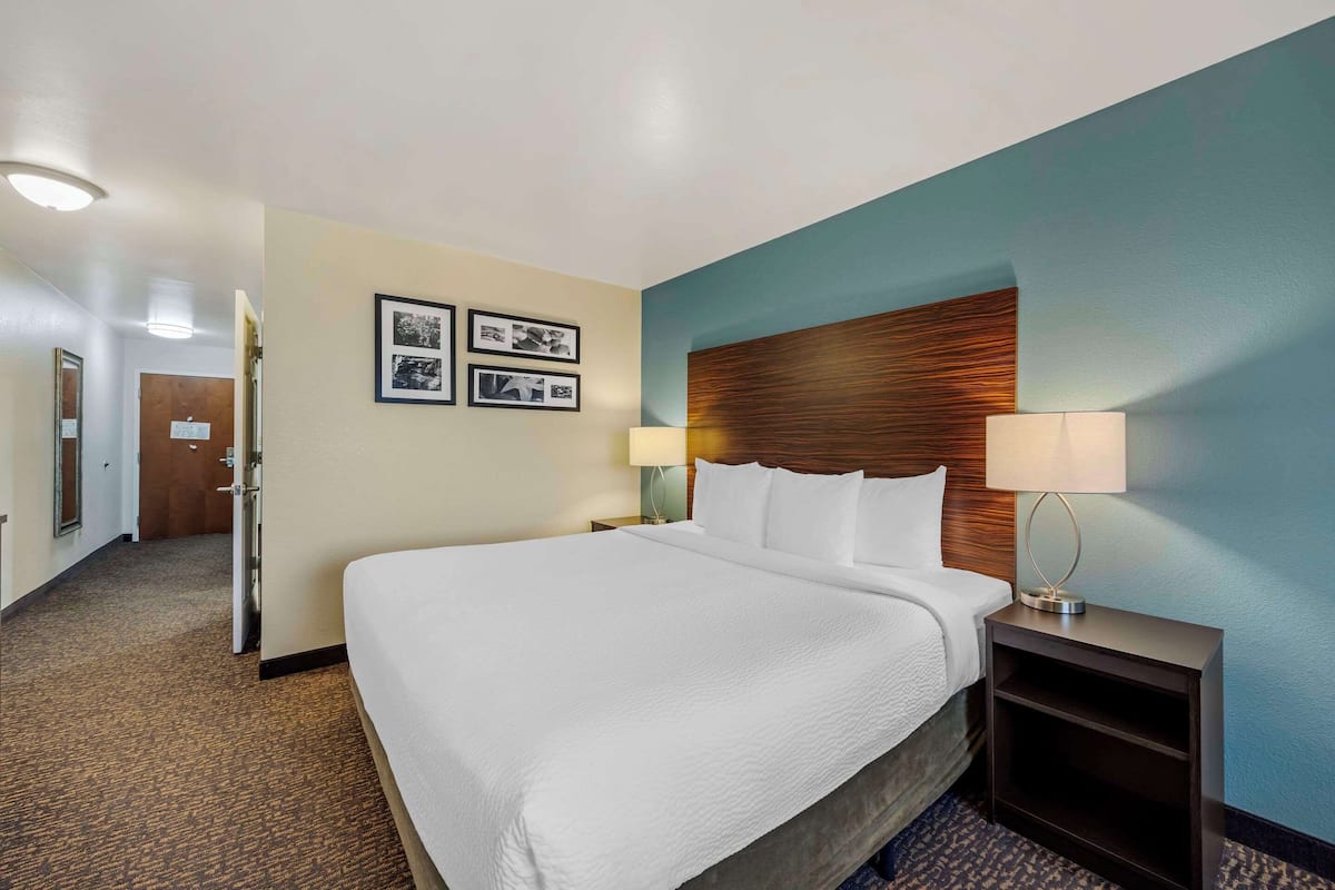 A hotel room with a large bed, a wooden headboard, a nightstand with a lamp, and a door leading to another room.