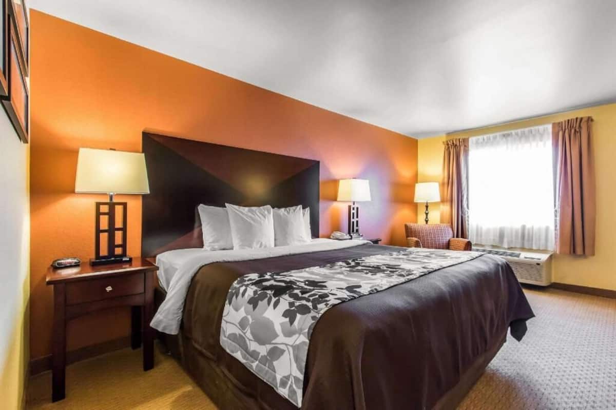 A hotel room with a large bed, two bedside tables, a window with curtains, and a wall-mounted air conditioning unit.