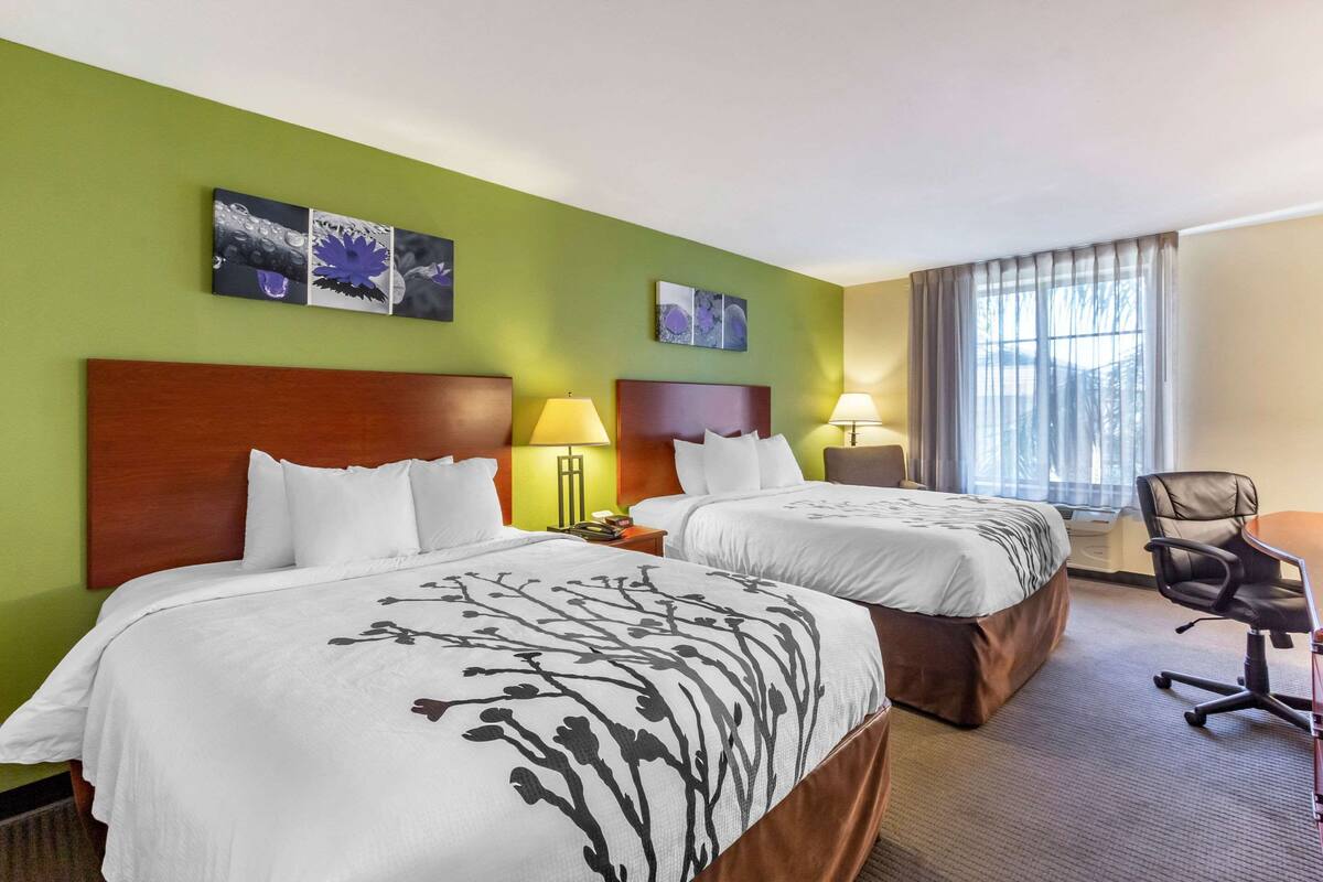 A hotel room with two beds, a desk, and a chair. There are wall art pieces above the beds and a window with curtains.