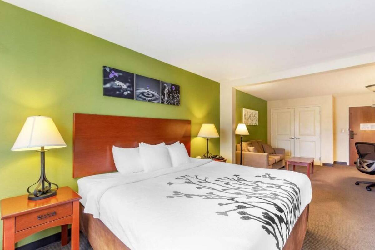 A hotel room with a bed, two bedside lamps, a nightstand, a chair, a desk, and a door.