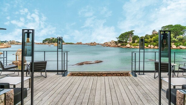 Una terraza de madera con asientos exteriores y vistas a una masa de agua tranquila, con rocas y vegetación al fondo.