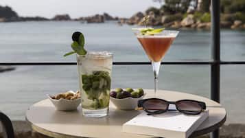 A table with two drinks, a bowl of nuts, a bowl of olives, and a pair of glasses on a seaside table.