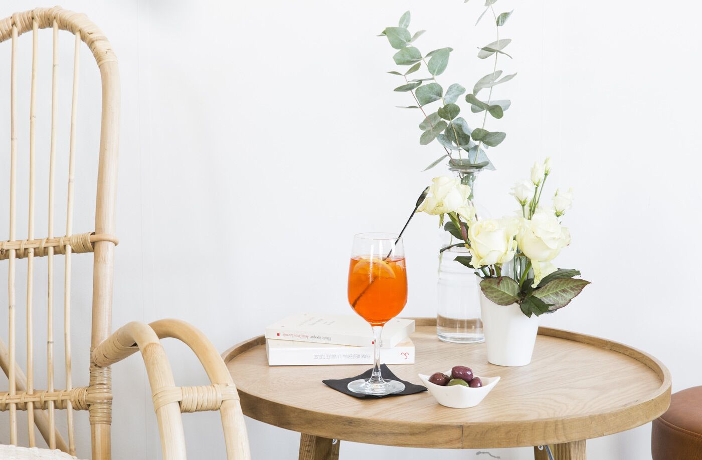 A round wooden table with a glass of orange juice, a book, a vase with white flowers, and a bowl of olives.