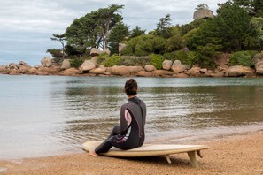 A person sitting on a surfboard on a pebble beach with a rocky shoreline and trees in the background. - Le Castel Beau Site (Perros-Guirec)