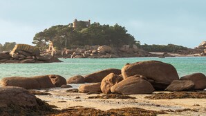Un paisaje costero con grandes rocas, una playa de arena y una estructura similar a un castillo en una colina.