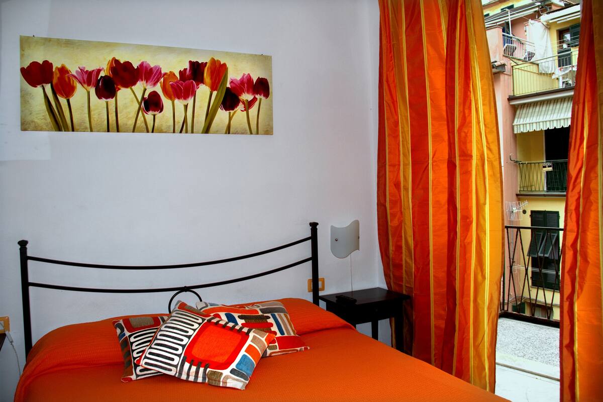 A bed with an orange bedspread and pillows, a painting of tulips on the wall, and a view of a balcony with orange curtains.